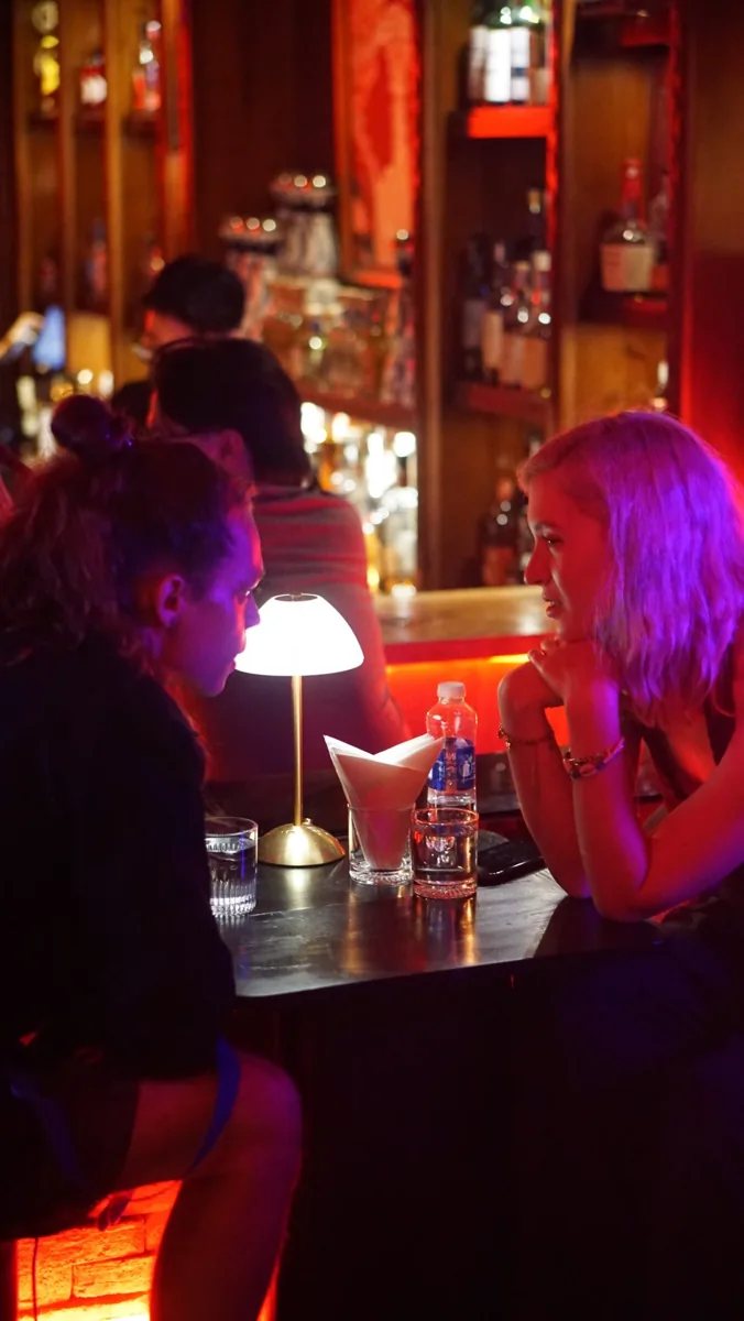Guests at Her Room cocktail bar under red and purple lighting; the shared programme advertises daily happy hour, Friday Bestie Room and Friday–Saturday DJ nights.