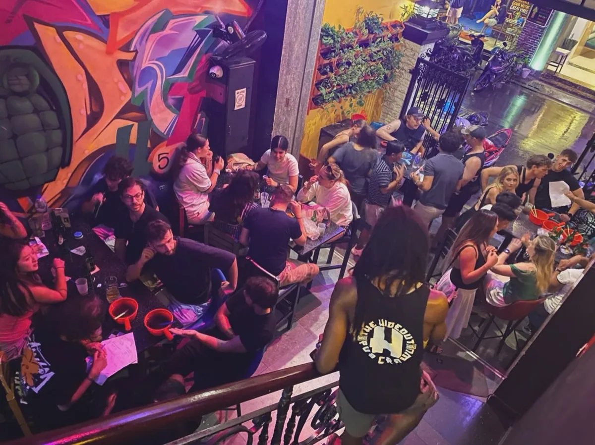 Guests at tables in The Hideout Hostel Saigon social bar, seen from the staircase beside a colourful wall mural.