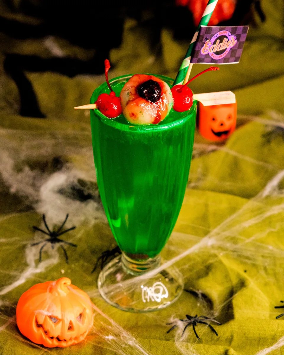 Green 'Witches Brew' mocktail with eyeball garnish and cherries in a tall glass, Eddie's Halloween Week branded flag, Halloween spiderwebs and pumpkin decorations